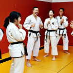 A diverse group of people (20s to 50s) in taekwondo uniforms, sweating and laughing together during a training session. Some are helping each other with techniques. The atmosphere is friendly and supportive, showing camaraderie and a sense of community.